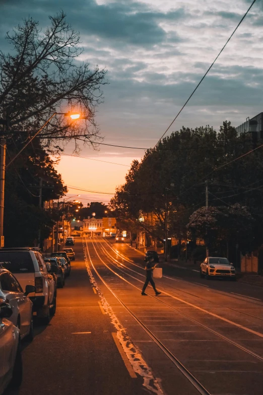 a person walking down a street next to parked cars, by Winona Nelson, unsplash contest winner, north melbourne street, sunset evening lighting, street tram, city lights made of lush trees