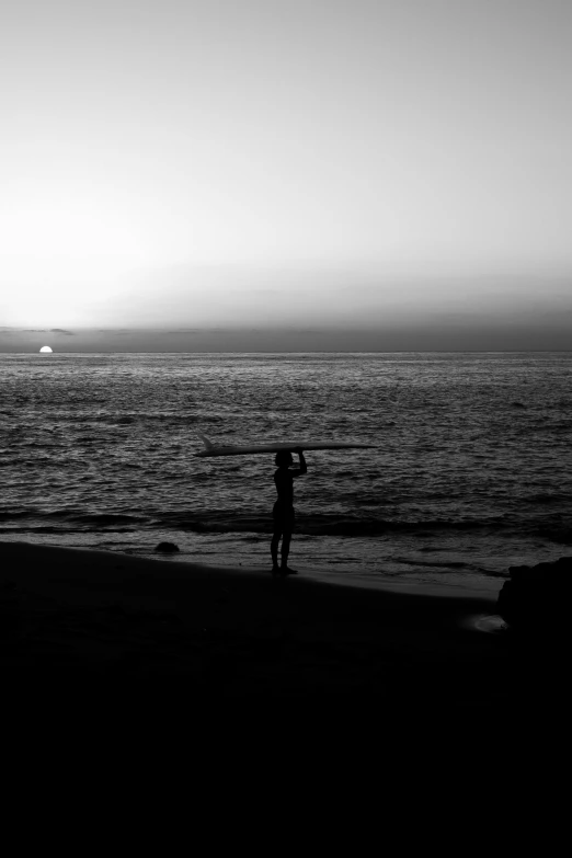 a man standing on top of a beach next to the ocean, a black and white photo, ((sunset)), paddle of water, :: morning, carson ellis