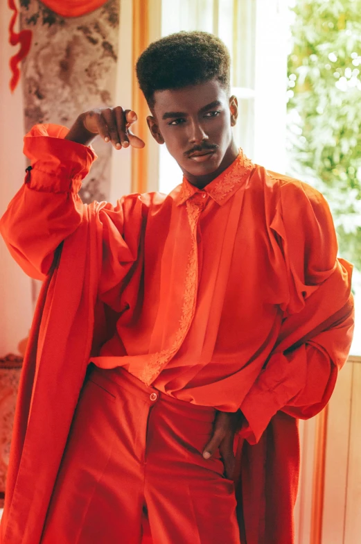 a man in an orange robe standing in front of a window, an album cover, inspired by Theo Constanté, trending on pexels, beautiful androgynous prince, red clothes, model posing, dark-skinned