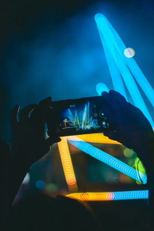 a person taking a picture with a cell phone, a hologram, pexels contest winner, interactive art, neon blue and yellow lights, concert lights, blue and orange rim lights, laser beam ; outdoor