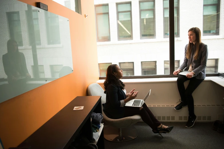 a couple of women sitting next to each other in a room, by Chris Rallis, unsplash, cubical meeting room office, gray and orange colours, next to window, wide high angle view