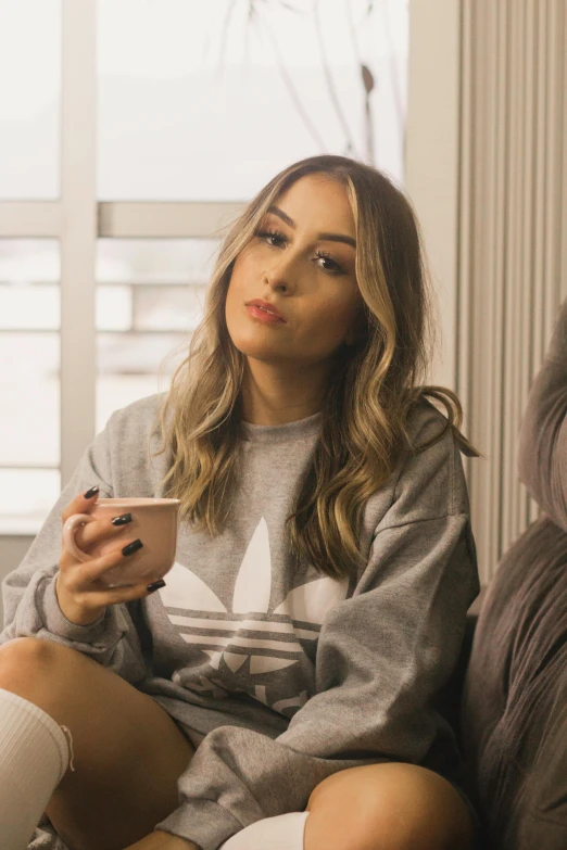 a woman sitting on a couch holding a cell phone, inspired by Julia Pishtar, trending on pexels, chloe bennet, wearing sweatshirt, ash blond greyish hair, with a white mug