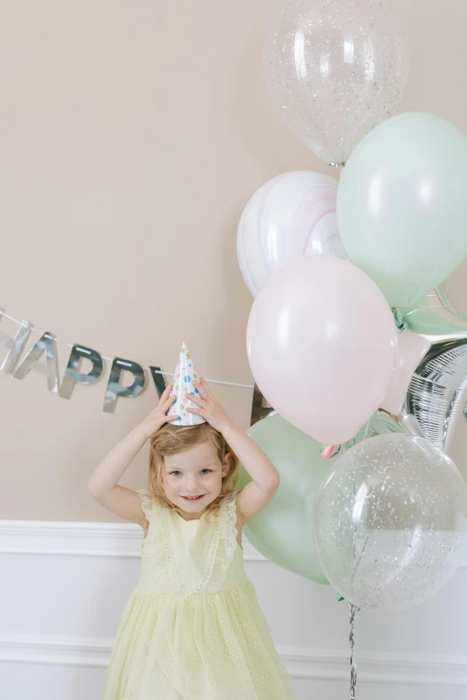 a little girl standing in front of a bunch of balloons, wearing a light grey crown, pastel color theme, smiling, full product shot