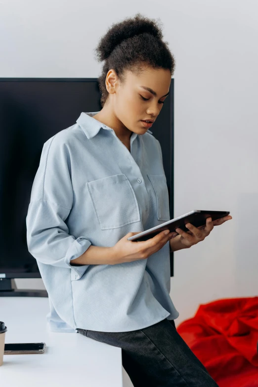 a woman standing in front of a tv holding a tablet, trending on pexels, wearing a light blue shirt, reading, androgynous person, tessa thompson inspired