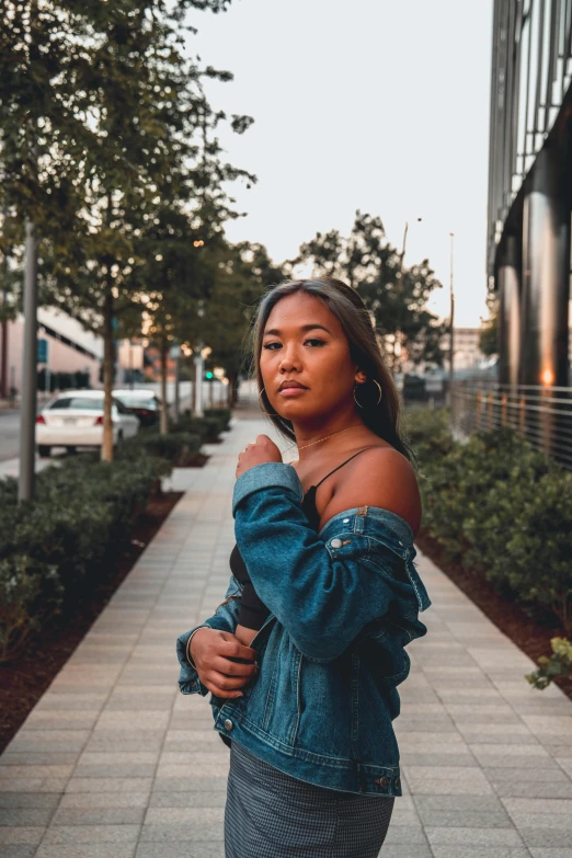 a woman standing on a sidewalk wearing a denim jacket, by Robbie Trevino, trending on unsplash, happening, portrait of modern darna, taken at golden hour, asian descent, slightly tanned