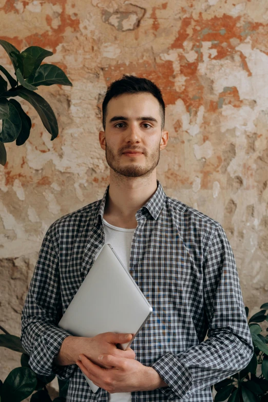 a man standing in front of a wall holding a laptop, by Adam Marczyński, wearing a light shirt, 21 years old, profile image, multiple stories