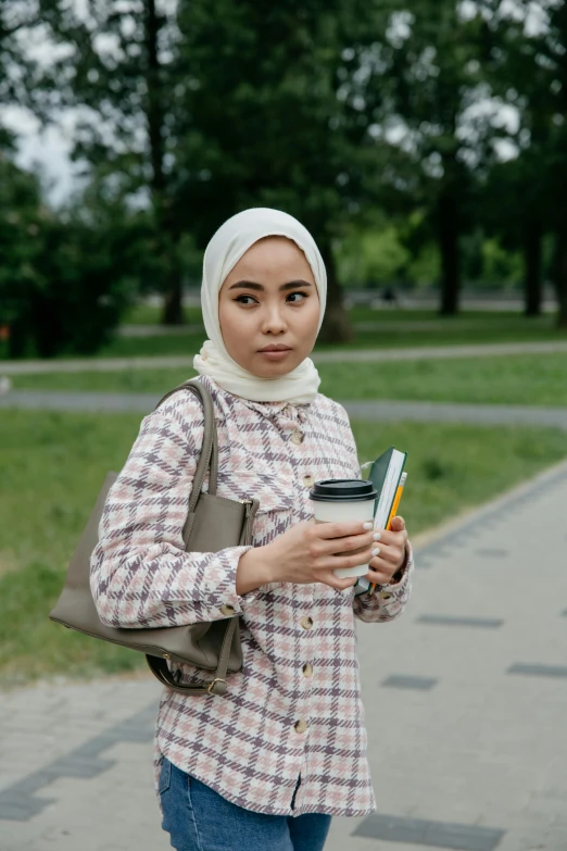 a woman standing on a sidewalk holding a cup of coffee, an album cover, inspired by Maryam Hashemi, trending on unsplash, hyperrealism, holding books, malaysian, at a park, concerned expression