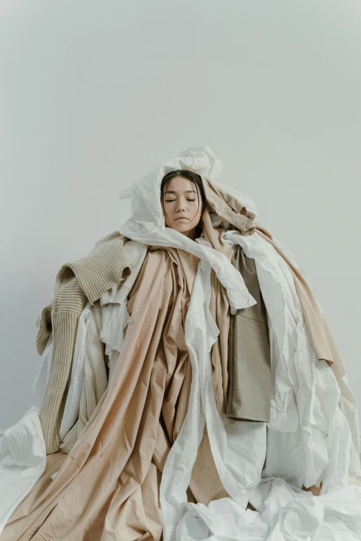 a woman sitting on a bed covered in a blanket, inspired by Anna Füssli, unsplash contest winner, conceptual art, pile of bodies, multilayered outfit, cardboard, neutral colours