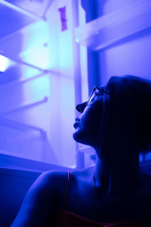 a woman sitting in front of an open refrigerator, pexels contest winner, light and space, blue and purle lighting, looking upwards, sunbathed skin, blue bioluminescent plastics