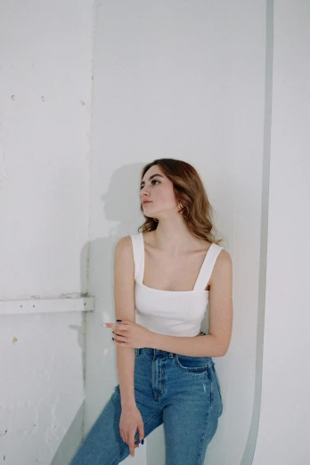 a woman in a white tank top leaning against a wall, inspired by Elsa Bleda, trending on pexels, realism, soft hair, in a white room, square jaw-line, full body picture