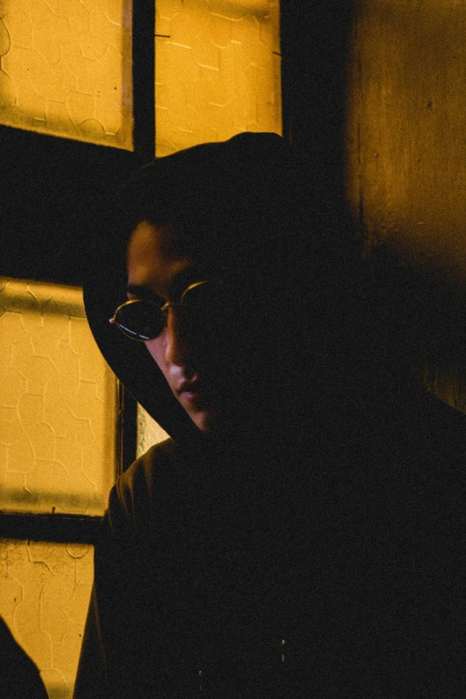 a man in a hoodie standing in front of a window, inspired by Bernardino Mei, unsplash, serial art, wearing gold glasses, korean film noir, female image in shadow, night photo
