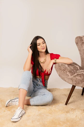 a woman sitting on the floor next to a chair, by Julia Pishtar, trending on pexels, arabesque, long hair and red shirt, dua lipa, jeans, avatar image