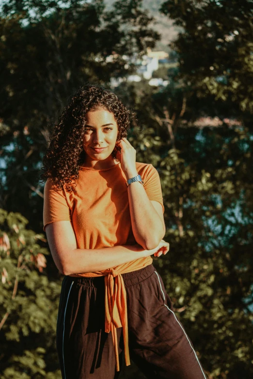 a woman standing in front of trees talking on a cell phone, short golden curls, wearing an orange t shirt, tanned ameera al taweel, wearing a black t-shirt