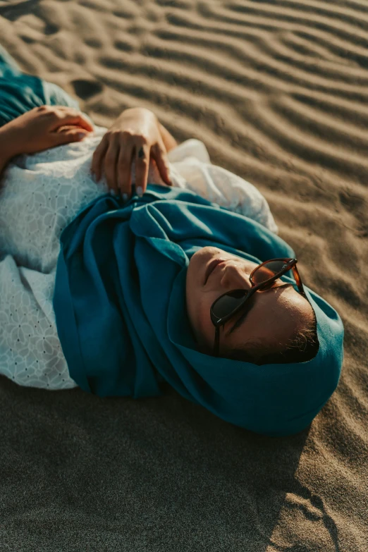 a woman laying on top of a sandy beach, unsplash contest winner, hurufiyya, hijab, cold shades, teal, wearing simple robes