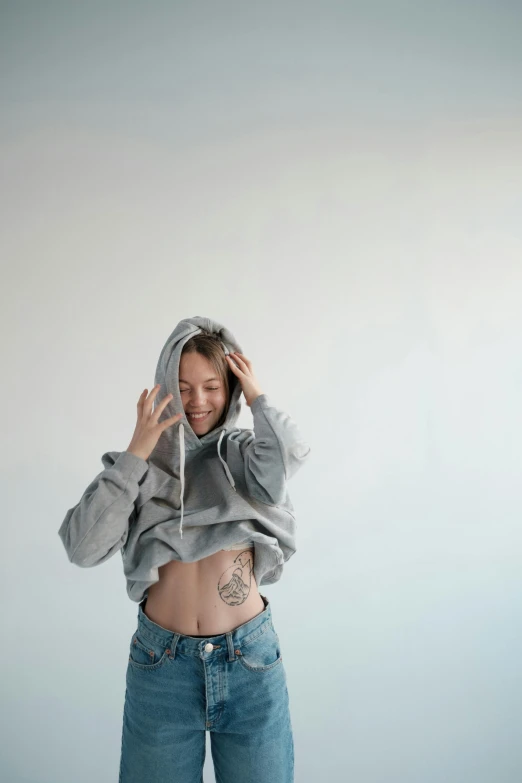 a woman in a hoodie standing with her hands on her head, a tattoo, trending on pexels, hyperrealism, exposed midriff, light grey backdrop, baggy jeans, gif