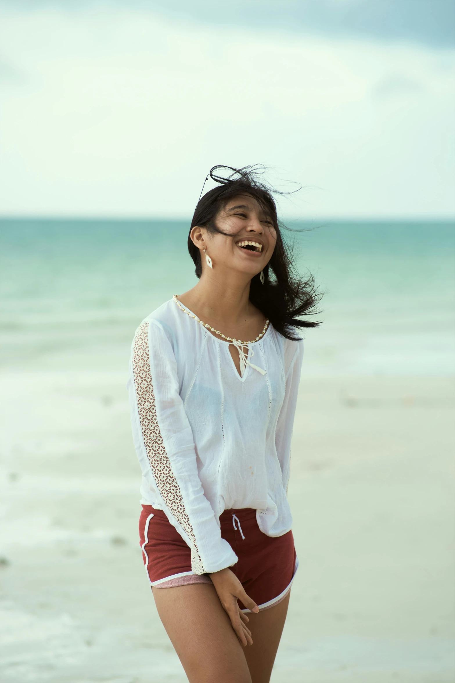 a woman standing on a beach next to the ocean, earing a shirt laughing, malaysian, white sleeves, promo photo