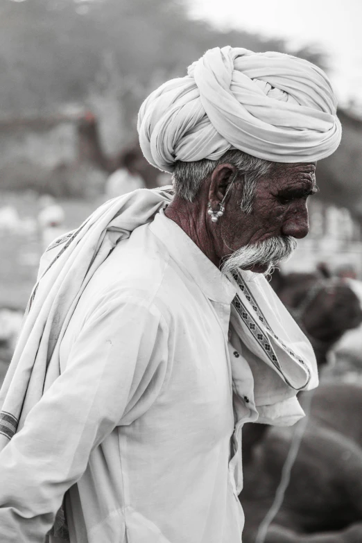 a man standing in front of a herd of cows, a black and white photo, inspired by Steve McCurry, pexels contest winner, wearing a turban, white grey color palette, vendors, close - up profile face