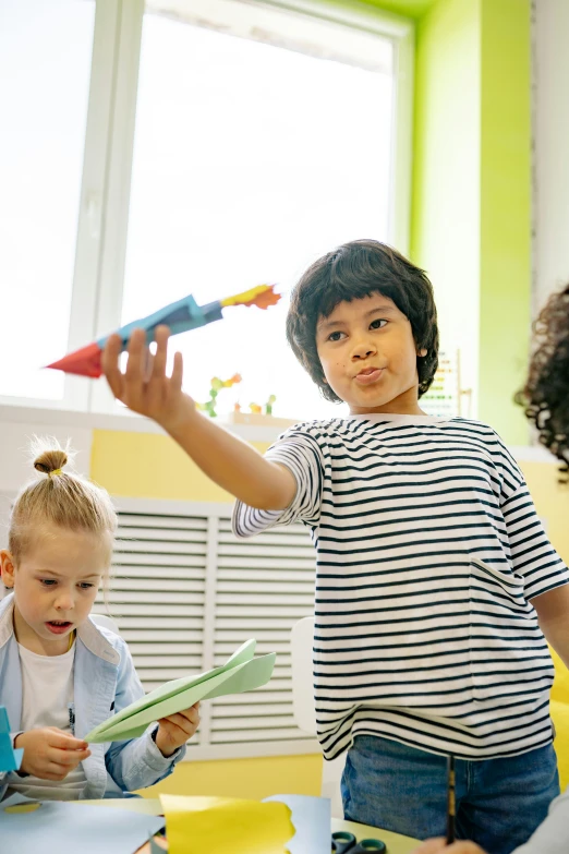 a group of children sitting around a table, holding a rocket, standing in class, promo image, playful creativity