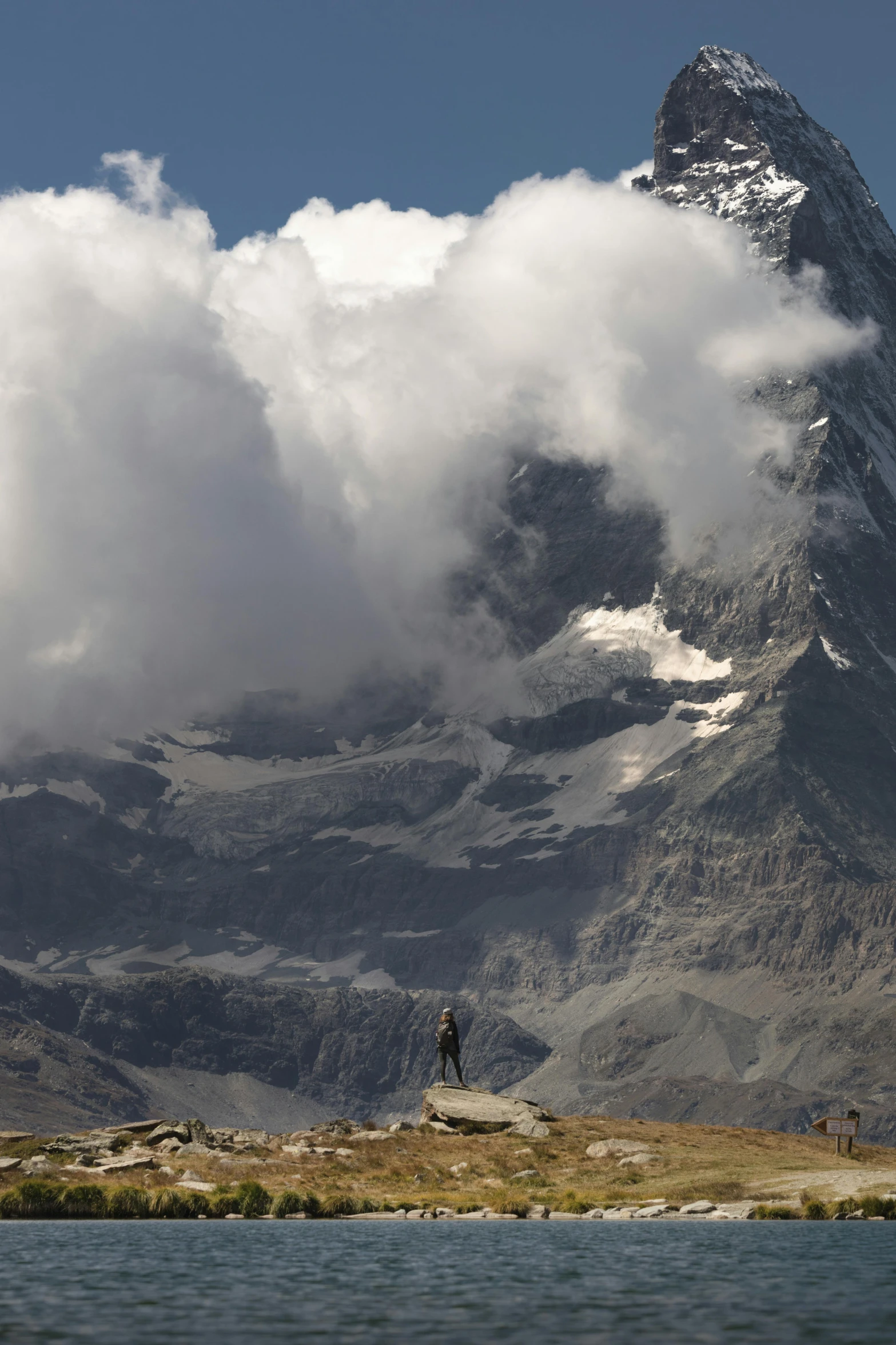 a man standing on top of a mountain next to a body of water, a matte painting, by Peter Churcher, trending on unsplash, swiss alps, towering cumulonimbus clouds, national geograph, standing atop a pile of rubble