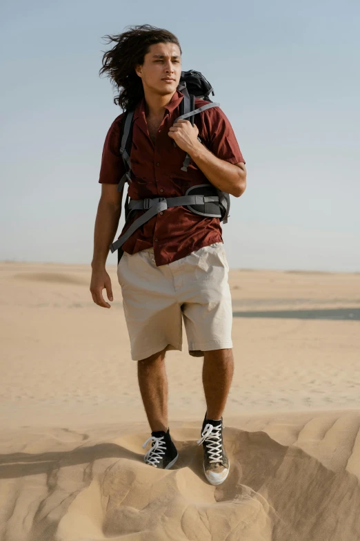 a man standing on top of a sand dune, a portrait, trending on pexels, cargo shorts, with a backpack, red shirt brown pants, arab man