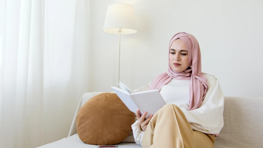 a woman sitting on a couch reading a book, hurufiyya, white and pink, wearing a head scarf, professional image, very calm and wholesome