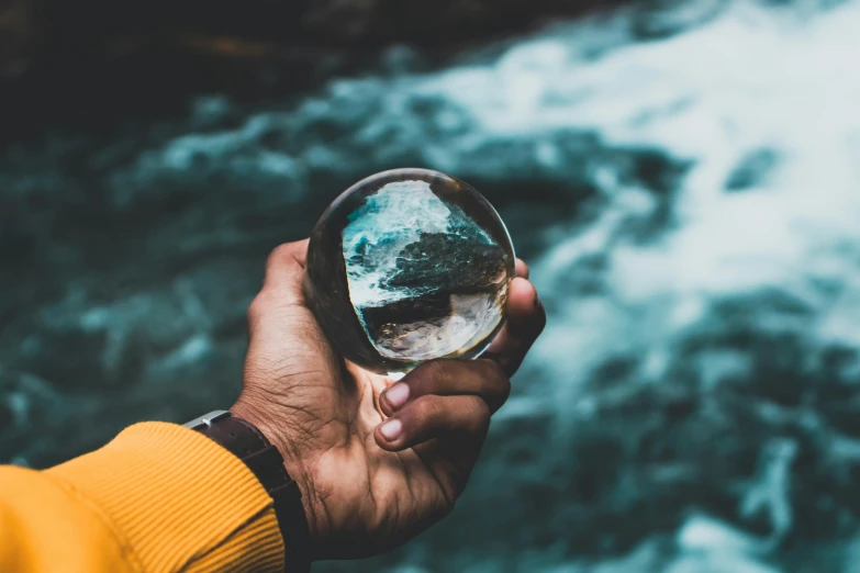 a person holding a glass in front of a river, by Christen Dalsgaard, pexels contest winner, holding a crystal ball, glacier, flatlay, magnified