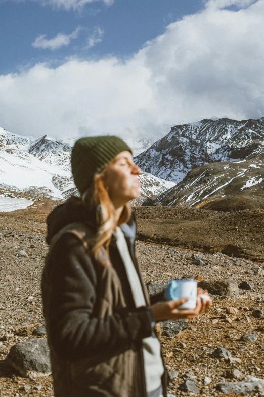 a woman standing in front of a mountain with a cup of coffee, by Jacob Toorenvliet, trending on unsplash, drinking cough syrup, with black beanie on head, andes, icon