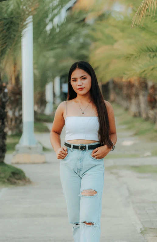 a woman standing on a sidewalk next to a palm tree, by Robbie Trevino, trending on pexels, croptop, south east asian with round face, teenage girl, wearing : tanktop