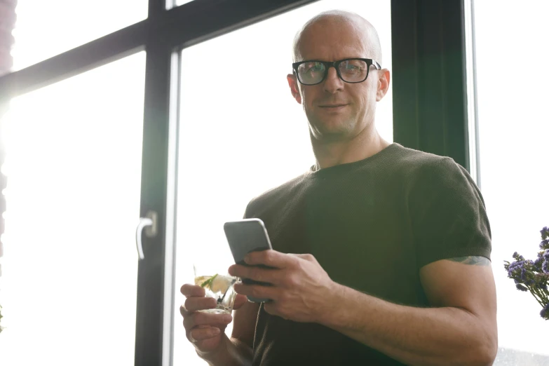 a man standing in front of a window holding a cell phone, hairless, avatar image, wearing reading glasses, markus vogt