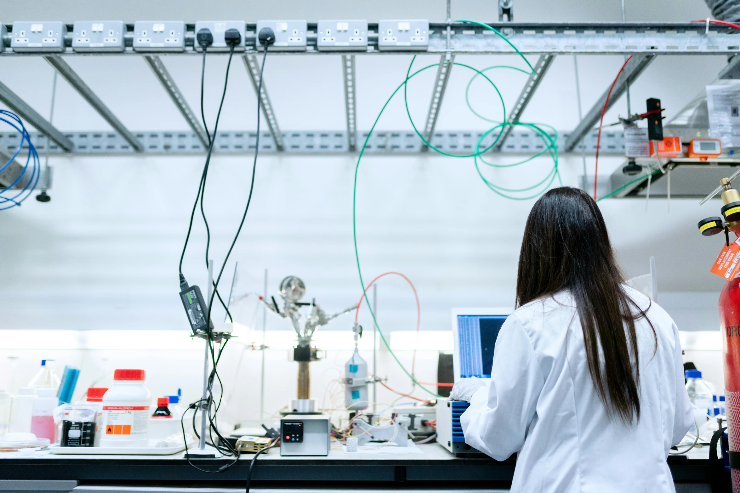 a woman in a lab coat standing in front of a counter, unsplash, cables and tubes, avatar image, back towards camera, liquid cooling