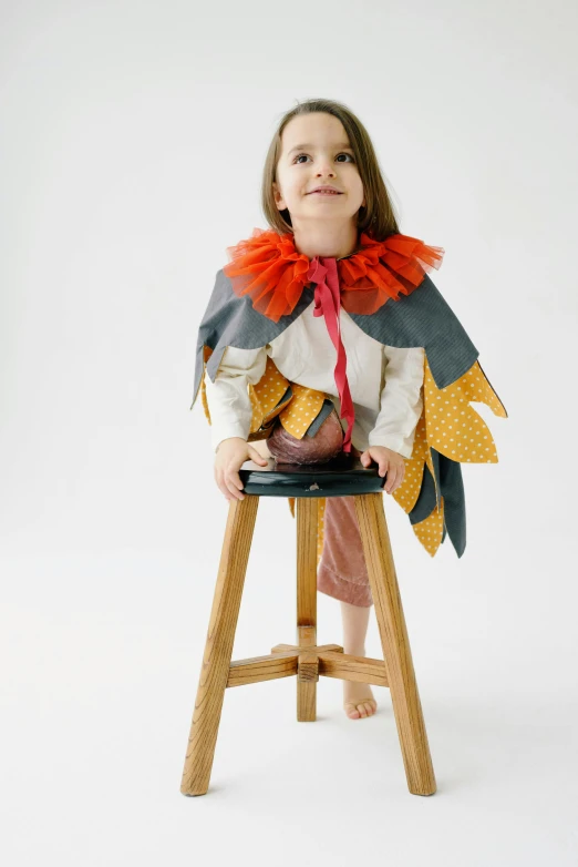 a little girl sitting on top of a wooden stool, wearing a cape, avian-inspired, grey orange, chicken