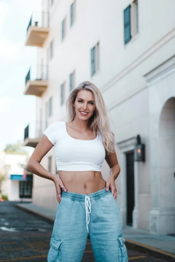 a woman standing in the middle of a street, by Robbie Trevino, crop shirt and strong abs, blonde and attractive features, in front of white back drop, dramatic smile pose