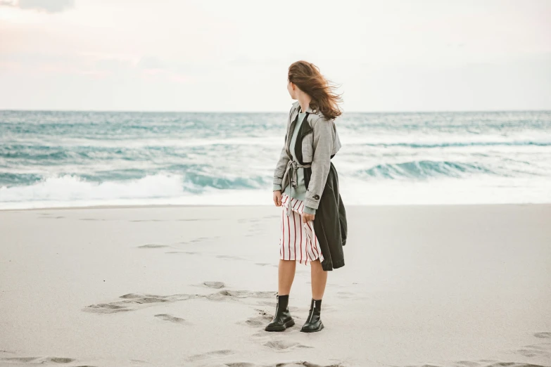 a woman standing on a beach next to the ocean, short skirt and a long jacket, portrait featured on unsplash, striped, looking at the ground