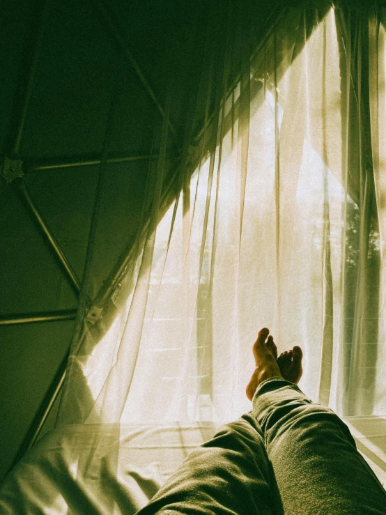 a person laying on a bed in front of a window, inspired by Elsa Bleda, unsplash contest winner, light and space, geodesic dome, glamping, green ambient light, mid view from below her feet
