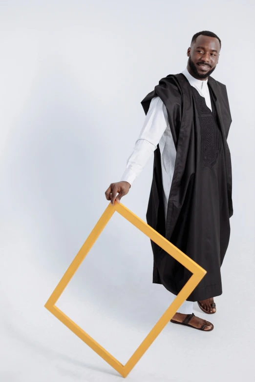 a man in a black robe holding a yellow frame, inspired by Ras Akyem, avant designer uniform, geometrical, 2019 trending photo, dressed thobe