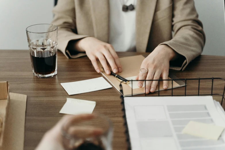 a woman sitting at a desk writing on a piece of paper, trending on unsplash, wearing a worn out brown suit, holding a drink, business meeting, tabletop game board