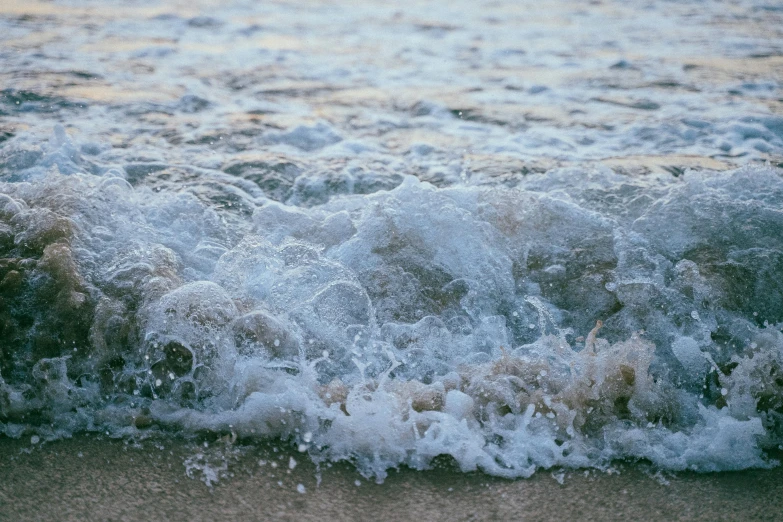 a large body of water sitting on top of a sandy beach, an album cover, inspired by Elsa Bleda, unsplash, renaissance, wave of water particles, close up shot, early evening, foamy waves