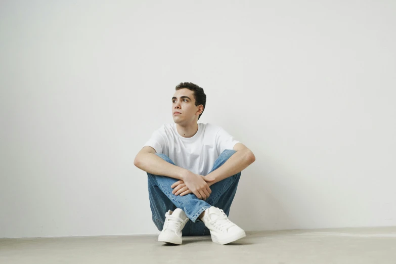 a man sitting on the floor in front of a white wall, inspired by Jean Malouel, pexels contest winner, hyperrealism, dressed in a white t shirt, declan mckenna, high soles, looking off into the distance