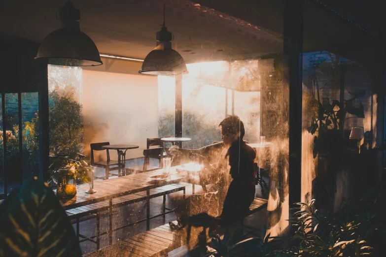 a person standing next to a table in a room, inspired by Elsa Bleda, pexels contest winner, light and space, raining outside the cafe, autumn sunrise warm light, misting, intense smoldering