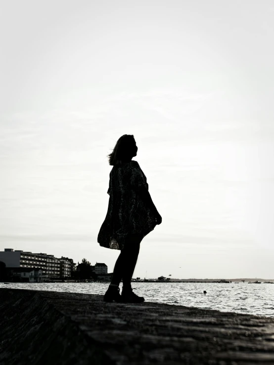 a woman standing on a beach next to a body of water, a black and white photo, by Christen Dalsgaard, unsplash, silhouette :7, wandering in the city, profile image, wearing a cape