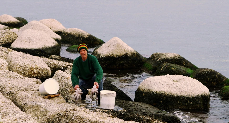 a man sitting on a rock next to a body of water, shells and barnacles, 2000s photo, tv still, filleting technique