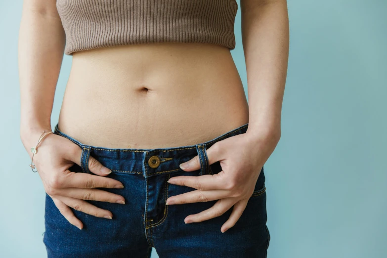 a close up of a woman's stomach with her hands on her hips, by Nicolette Macnamara, trending on pexels, relaxed. blue background, manuka, ( ( ( wearing jeans ) ) ), dry skin