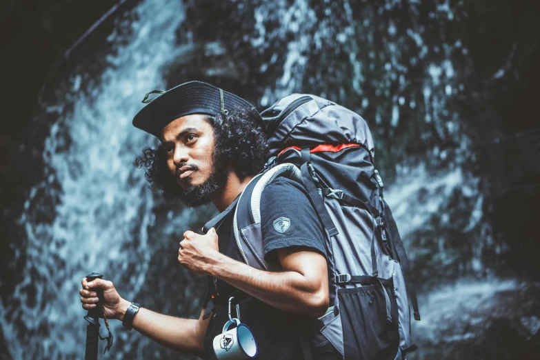 a man with a backpack standing in front of a waterfall, pexels contest winner, a black man with long curly hair, wearing adventure gear, outlive streetwear collection, malaysian