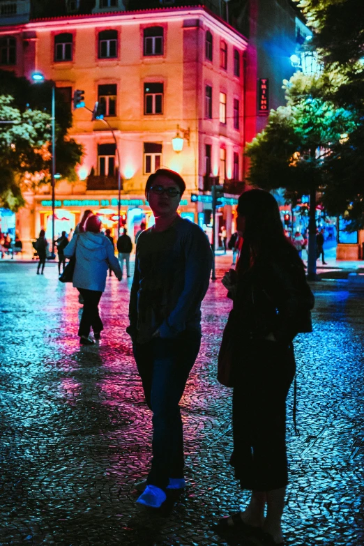 a group of people walking down a street at night, lisbon city at night, bisexual lighting, square, two people