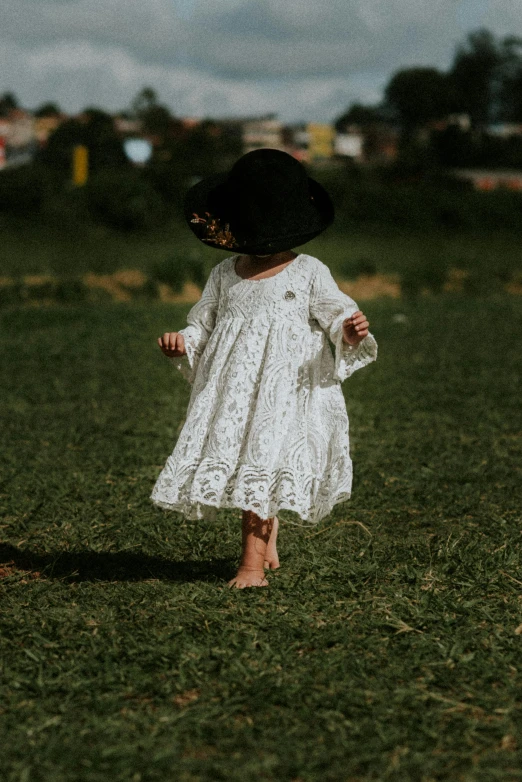 a little girl in a white dress and a black hat, inspired by Kate Greenaway, pexels contest winner, arabesque, walking on grass, dressed in a frilly ((lace)), boho neutral colors, square