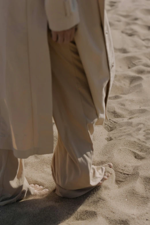 a couple of people standing on top of a sandy beach, an album cover, by Anna Boch, unsplash, renaissance, wearing a long beige trench coat, close-up on legs, large pants, detail texture