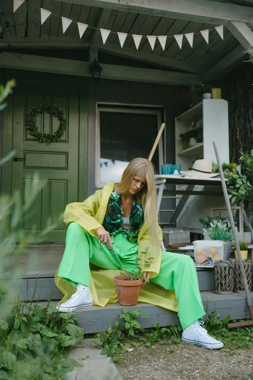 a woman sitting on a porch next to a potted plant, an album cover, trending on pexels, wearing green suit, long yellow hair, full body picture, wearing a duster coat