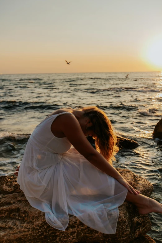 a woman in a white dress sitting on a rock by the ocean, pexels contest winner, romanticism, sunset glow around head, yoga pose, downward somber expression, mediterranean