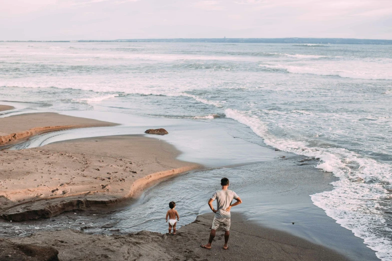 a man standing on top of a sandy beach next to the ocean, by Daniel Lieske, pexels contest winner, sumatraism, with a kid, currents, flatlay, big island