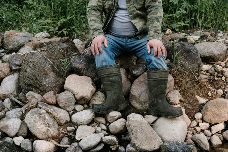 a man sitting on top of a pile of rocks, jeans and boots, swollen veins, dirty green clothes, family friendly